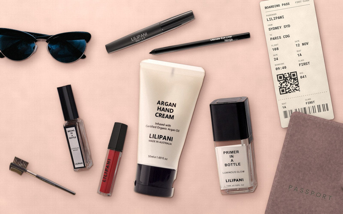 LILIPANI travel makeup flat lay with Argan Hand Cream, Primer In A Bottle, Brows in a Bottle, Lippy, mascara, eyeliner and a Sydney to Paris boarding pass on a soft blush pink background